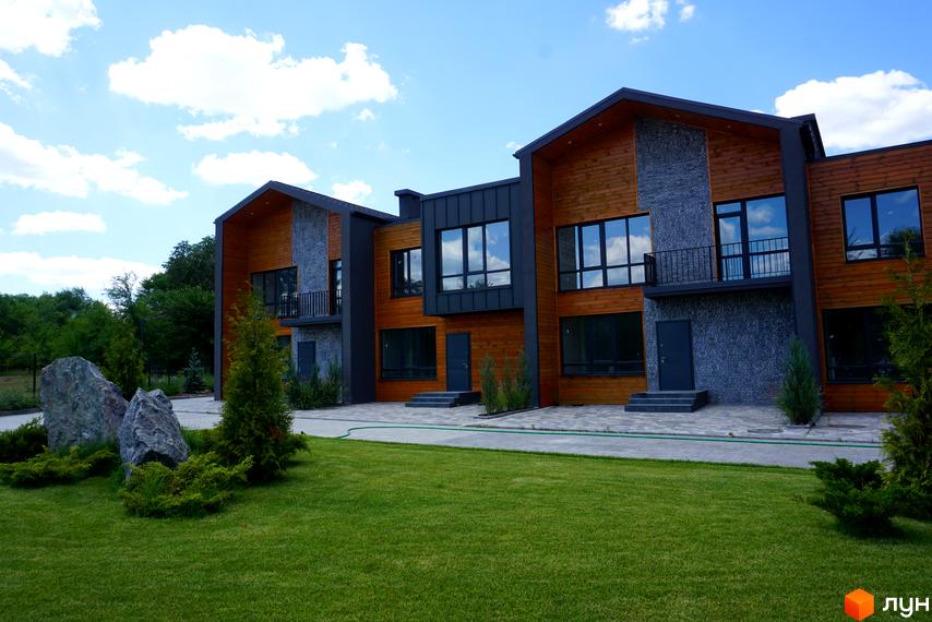 Котеджне містечко КМ ЛісоПаркове: фасади будинків з дерева та каменю, великі вікна, балкон, зелена галявина та декоративні камені.