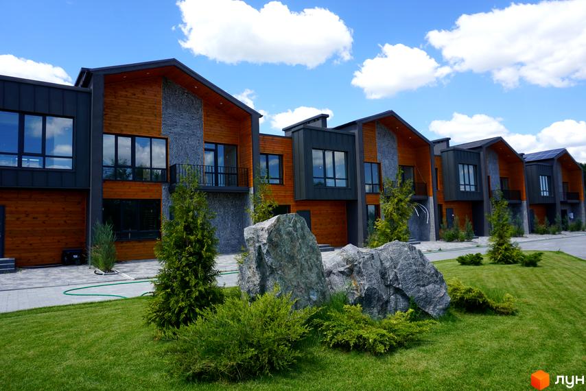 Котеджне містечко КМ ЛісоПаркове. Зовнішній вигляд будинків з дерев'яними та кам'яними фасадами, великими вікнами та прибудинковою територією з газоном і валунами.