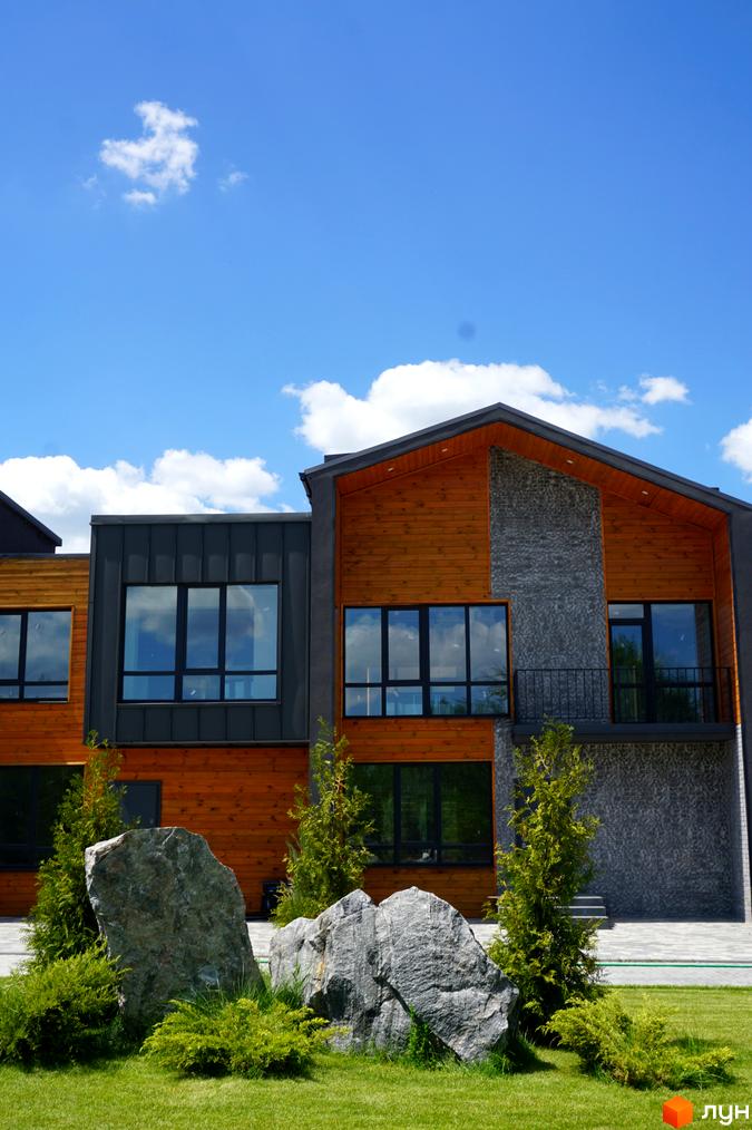 Котеджне містечко КМ ЛісоПаркове. Будинок з дерев'яною та кам'яною обробкою фасадів, великими вікнами та зеленою прибудинковою територією з валунами.