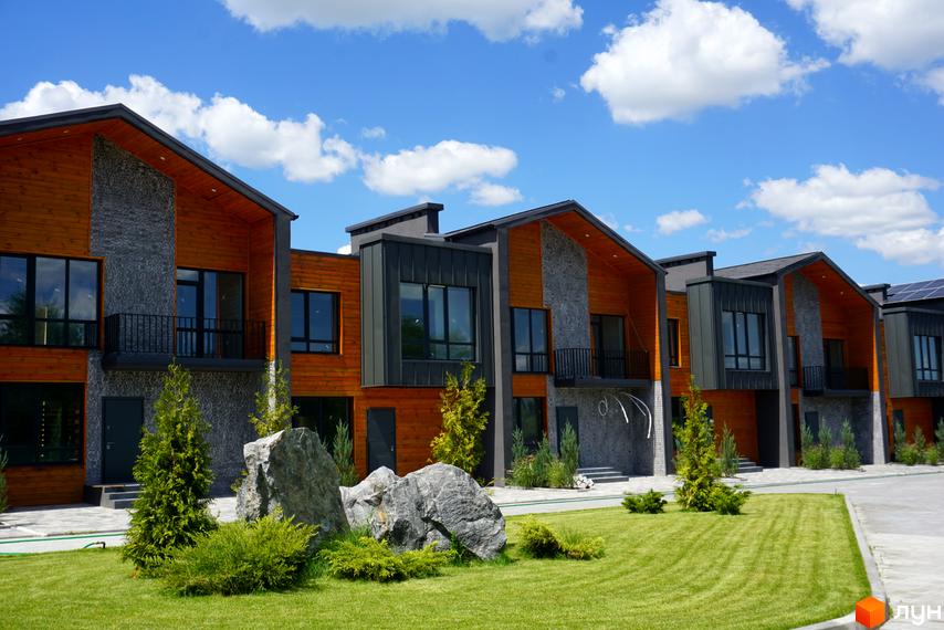 Котеджне містечко КМ ЛісоПаркове: фасади з дерева та каменю, зелена галявина з великим камінням та туями.