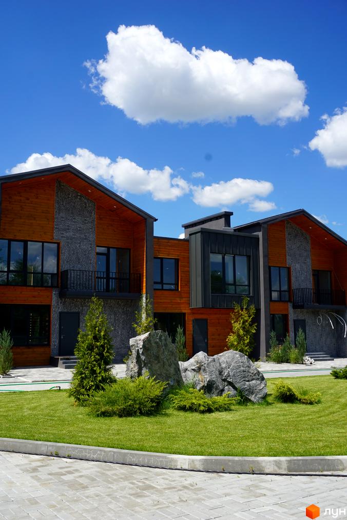 Котеджне містечко КМ ЛісоПаркове: двоповерхові будинки з дерев'яними та кам'яними фасадами, великими вікнами та доглянутою зеленою прибудинковою територією з декоративними каменями.