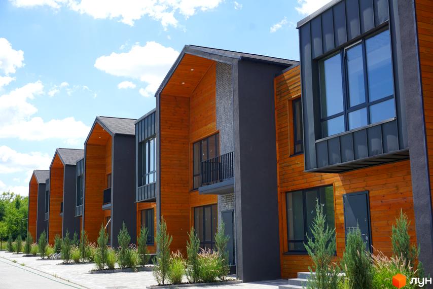 Котеджне містечко КМ ЛісоПаркове: фасади будинків з дерев'яним оздобленням та великими вікнами, зелені насадження перед будинками.