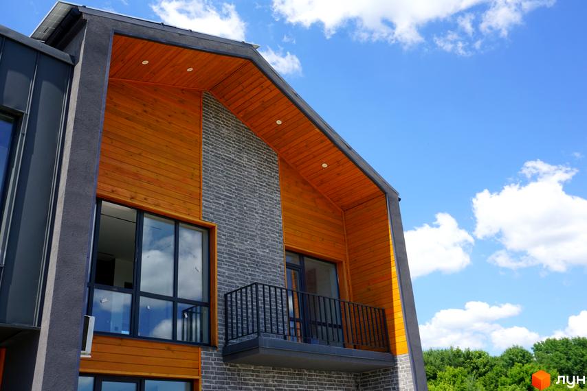 Зовнішній вигляд будинку житлового комплексу КМ ЛісоПаркове з дерев'яними елементами та сірою цегляною кладкою на фоні блакитного неба з хмарами. Балкон з чорною металевою огорожею.