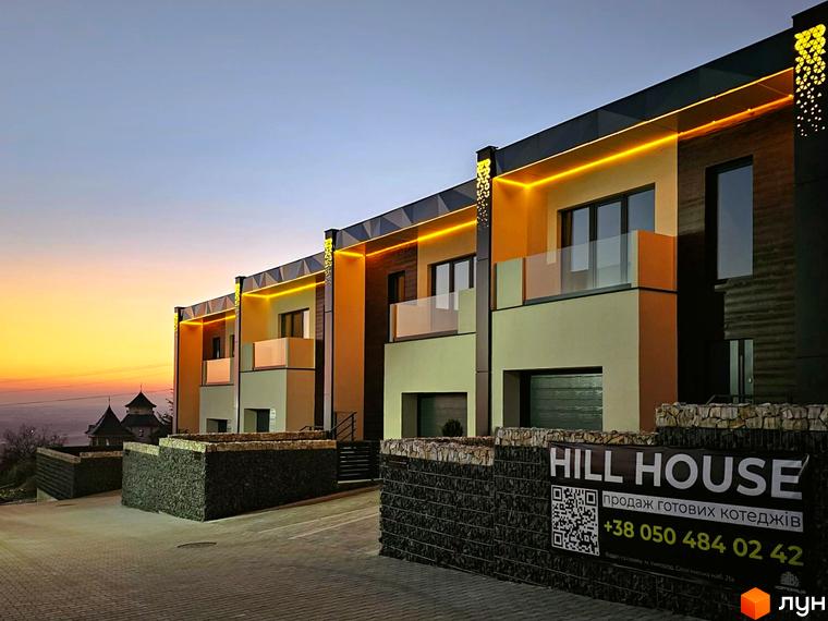 Таунхауси HILL HOUSE з жовтою підсвіткою та скляними балконами на фоні вечірнього неба. Територія перед будинками огороджена кам'яними парканами.