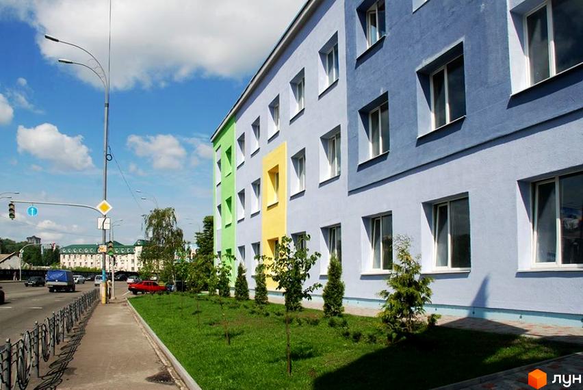 Новобудова Loft House Podol з яскравим фасадом у блакитних, зелених та жовтих кольорах. Поруч доглянута прибудинкова територія з газоном та молодими деревами.