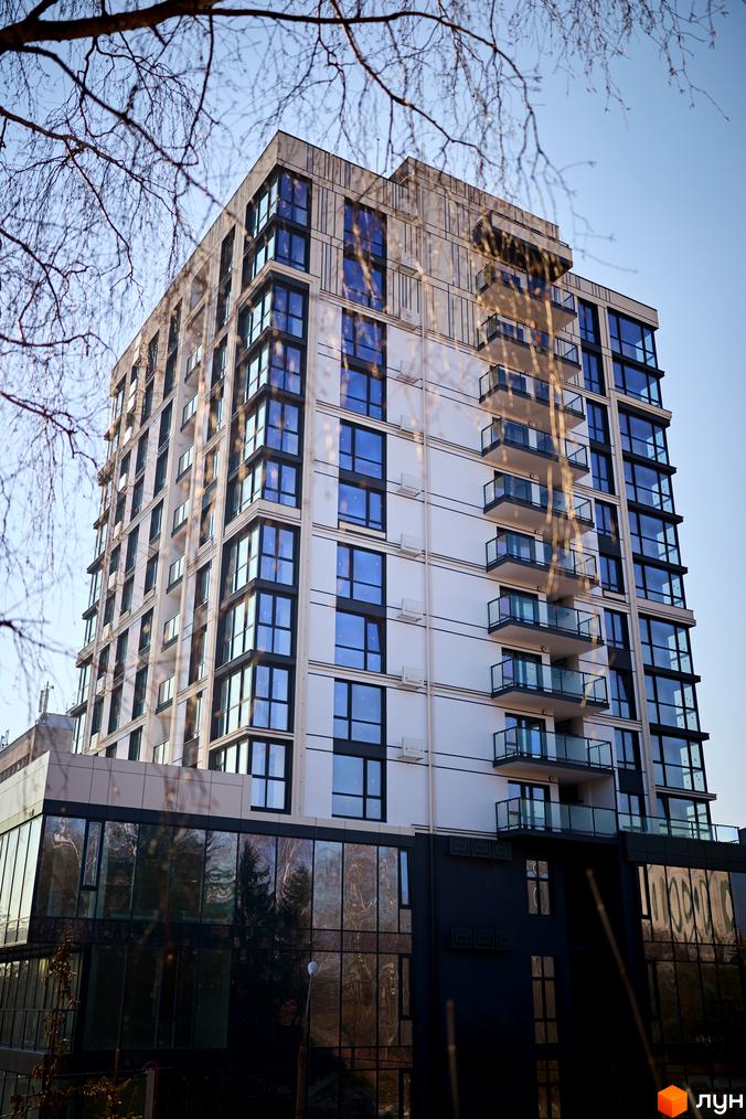 Зовнішній вигляд житлового комплексу ЖК SOURENGE: багатоповерхова будівля з панорамними вікнами та балконами на тлі блакитного неба. На першому поверсі розташовані комерційні приміщення з великими вікнами, що відбивають дерева.