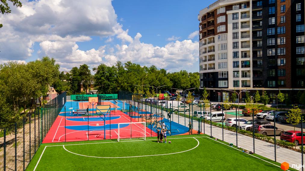 Висотна будівля житлового комплексу з фасадом у світлих тонах. На прибудинковій території спортивний майданчик з футбольним полем та баскетбольним кортом.
