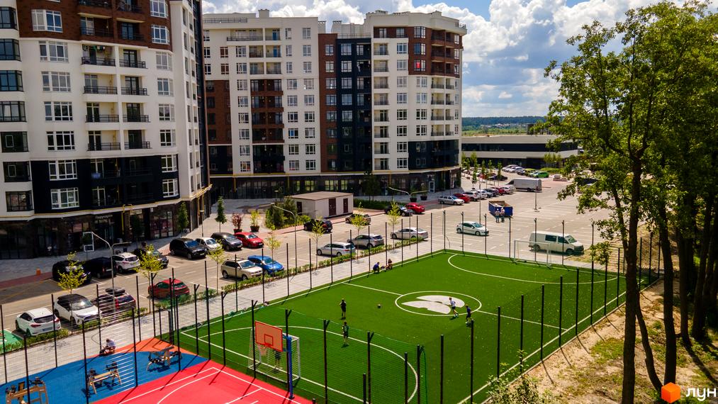 Високоповерховий будинок ЖК Синергія Сіті з великими вікнами, поруч спортивний майданчик та парковка для автомобілів.