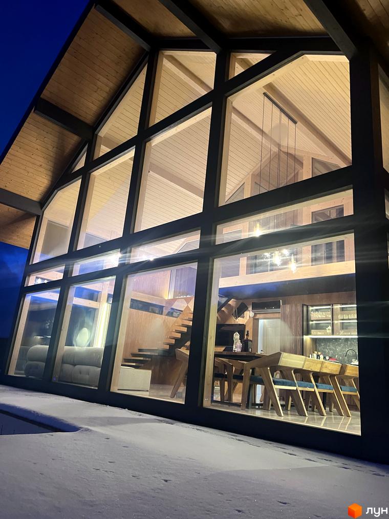 Будинок Котеджного містечка Шале Вище Неба з високим дерев'яним дахом та великими скляними фасадами, навколо якого лежить сніг.