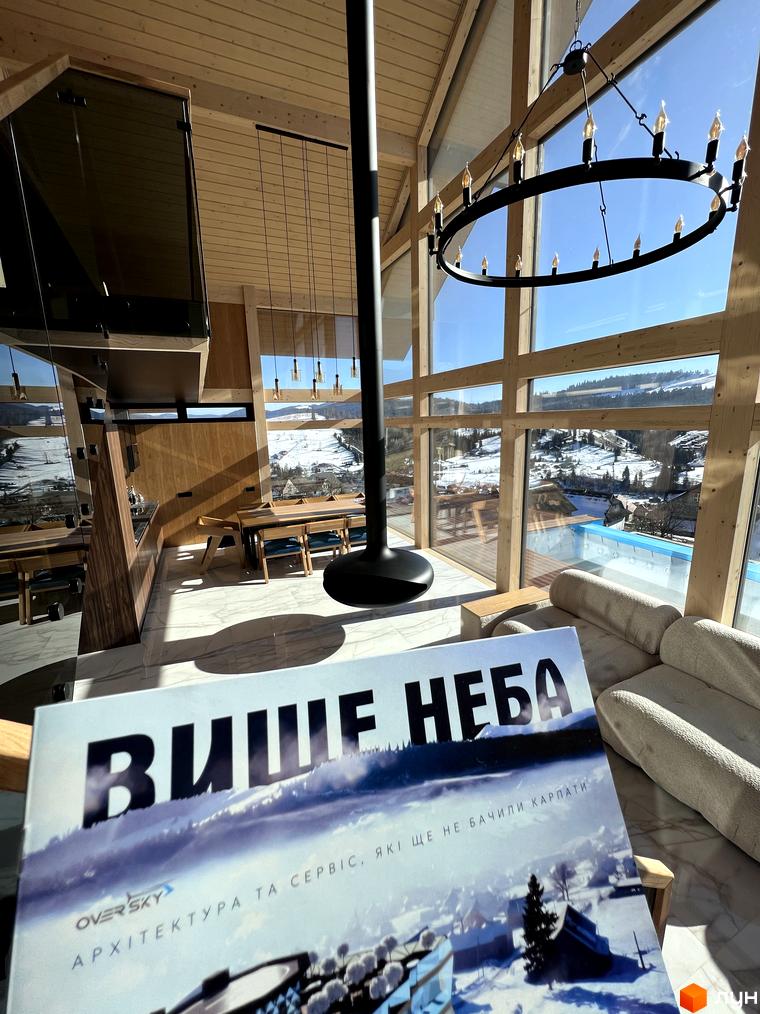 Панорамні вікна будинку Котеджного містечка Шале Вище Неба відкривають вид на засніжені гори. Видно дерев'яні конструкції та зовнішній басейн.