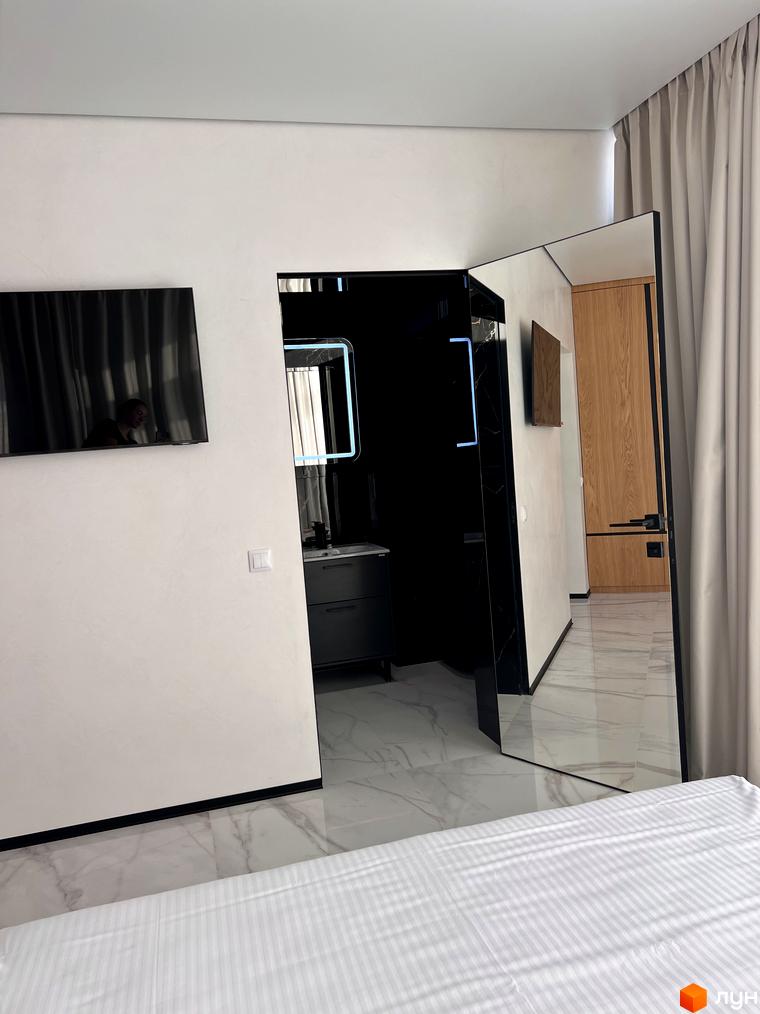 Простора кімната Котеджного містечка Шале Вище Неба з білими стінами, ліжком, телевізором та дзеркалом з підсвічуванням у ванній кімнаті.