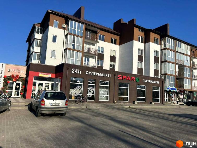 Зовнішній вигляд житлового комплексу Рать з фасадами білого та коричневого кольорів, вікнами з потрійним склінням та балконами. Поруч розташовані комерційні приміщення та парковка.