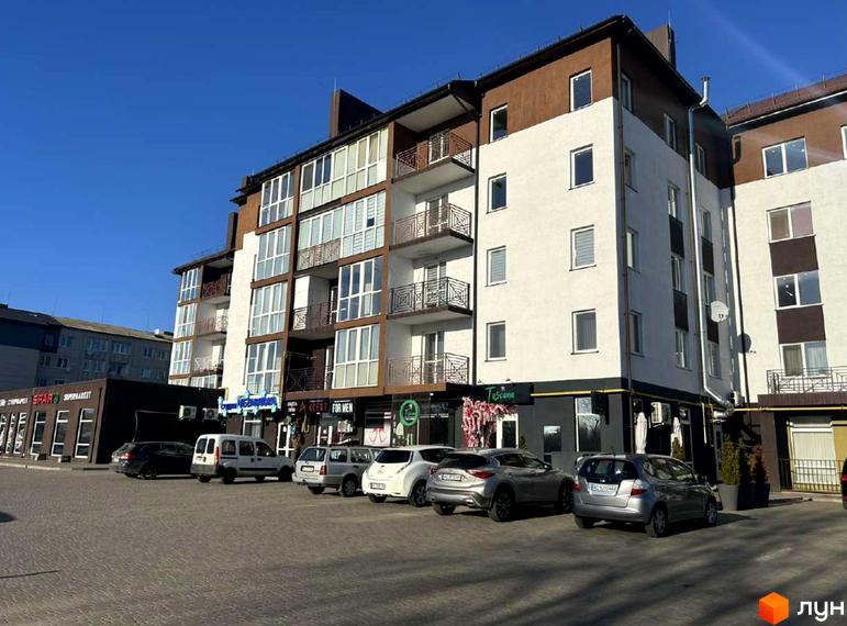 Зовнішній вигляд житлового комплексу Рать з фасадом білого та коричневого кольору, балконами та парковкою перед будинком.