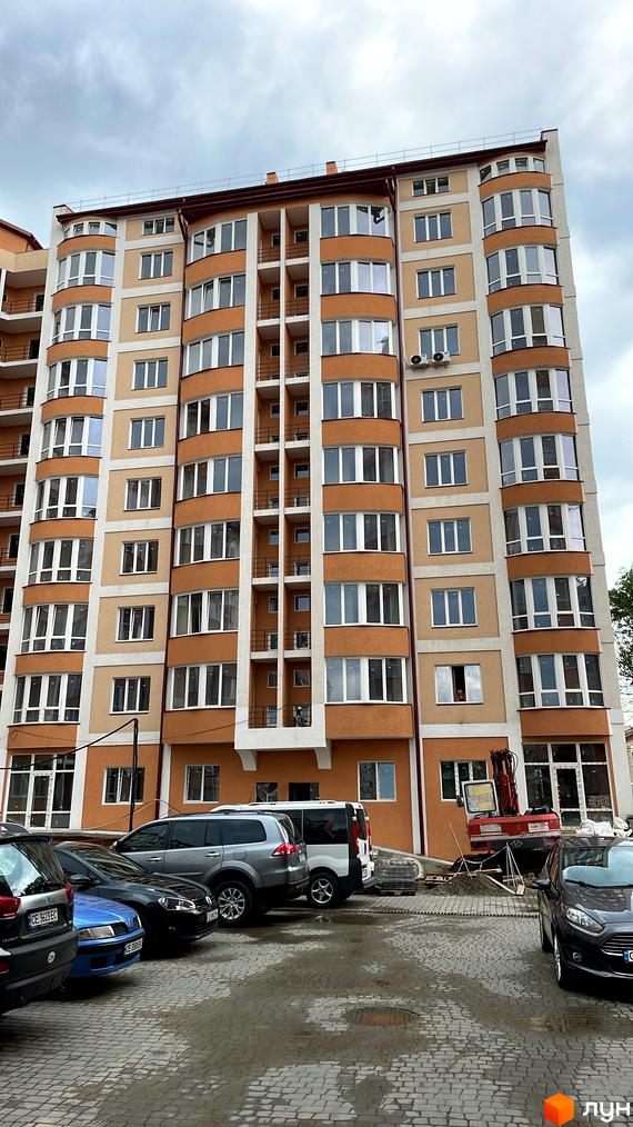 Зовнішній вигляд багатоповерхового будинку з коричневими та бежевими фасадами, великими вікнами та балконами. На прибудинковій території розташовані автомобілі та будівельна техніка.