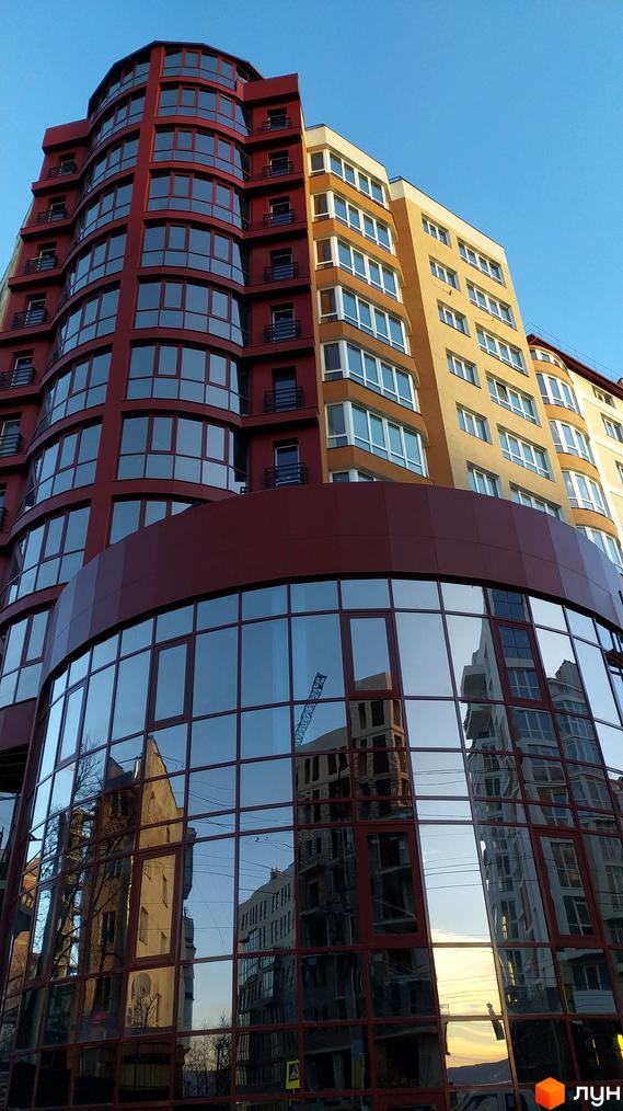 Фасад житлового комплексу на вул. Героїв Майдану, 53, з вигнутими червоними та жовтими секціями, великими панорамними вікнами та скляним цоколем, що відбиває сусідні будівлі та будівельний кран.