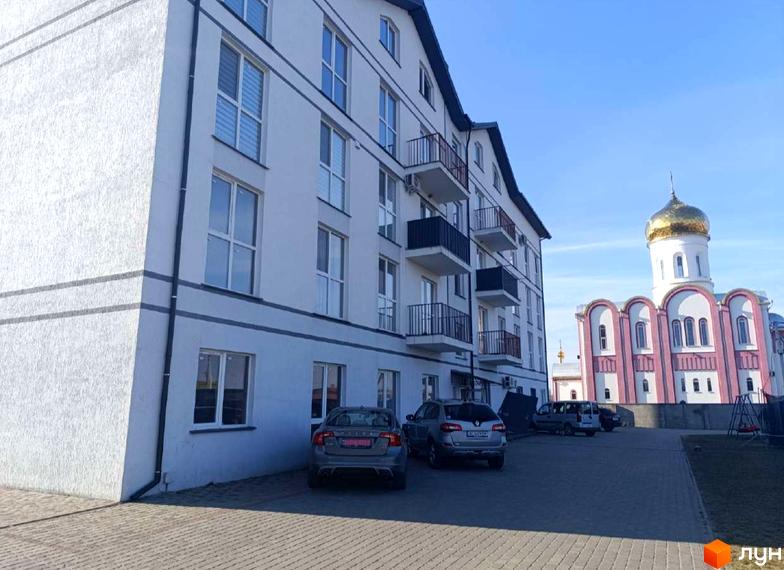 Біла багатоповерхівка з балконами та великими вікнами. На території біля будинку припарковані автомобілі, поруч церква з золотим куполом.