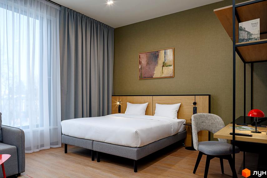 Інтер'єр спальні в апарт-комплексі. Ліжко з білою постільною білизною, стіл з лампою, стіни з картиною та шторами.