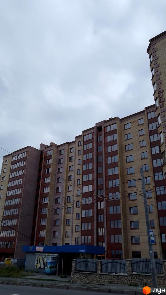 Багатоповерховий житловий комплекс на вулиці Леся Курбаса, з цегляним фасадом та великою кількістю вікон. Біля будинку розташована прибудинкова територія з парканом та зупинкою громадського транспорту.