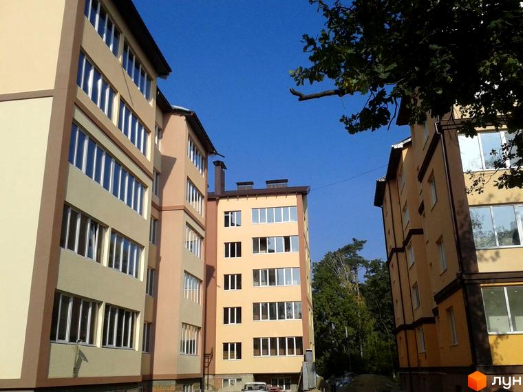 Фасад будівель Житлового комплексу Високий світлих відтінків з великими вікнами. Навколо новобудов багато зелених дерев та чисте блакитне небо.
