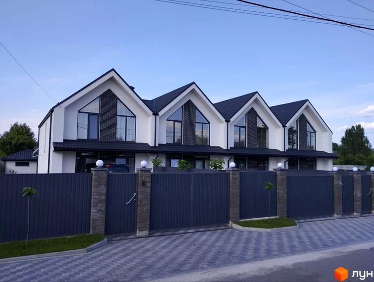 Три будинки з білими фасадами та чорними дахами, огороджені сірим парканом з кованими воротами. Дорога викладена бруківкою.