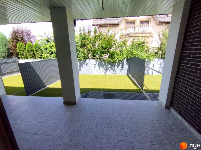 Вид на вхідну групу будинку з колонами, облицьованого цеглою. Навколо будинку газон, паркан та зелені насадження.