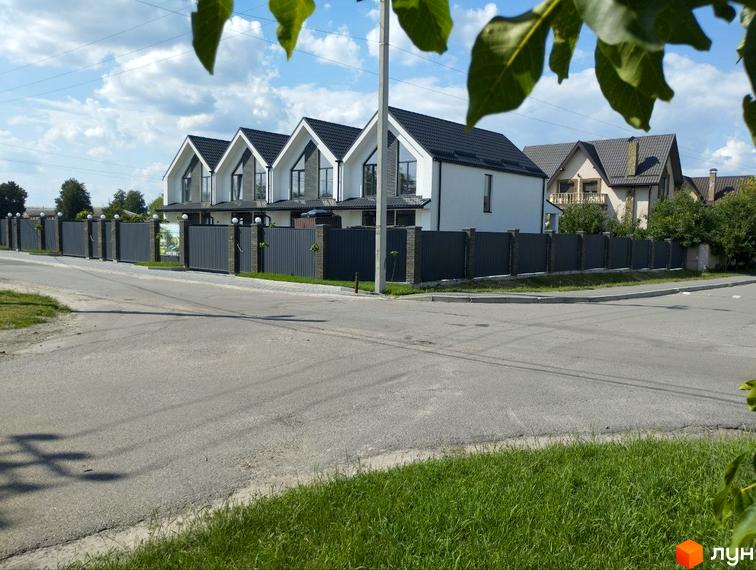 Триповерховий будинок з білими стінами та чорним дахом, огороджений сірим парканом. Навколо будинку асфальтована дорога та зелені насадження.