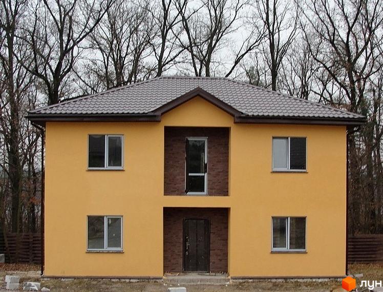 Котеджне містечко Круглик: двоповерховий будинок з жовтим фасадом, коричневим дахом та темними вікнами. Навколо видно дерева без листя та паркан.