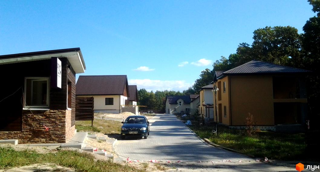 Котеджне містечко КМ Круглик: двоповерхові будинки з коричневими дахами та жовтими фасадами, розташовані вздовж бруківки. На території комплексу є зелені насадження та дерева.