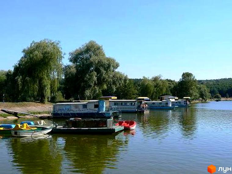 Котеджне містечко Круглик біля озера з човнами. На березі ростуть дерева, на воді видно кілька будинків на воді та катамарани.