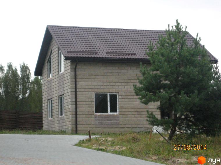 Котеджне містечко Круглик: двоповерховий будинок з коричневим дахом та сірими стінами з блоків, вікна з білими рамами. Навколо будинку зелені насадження та частина дороги.