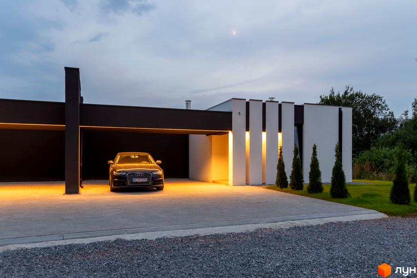 Сучасний будинок Duplex Comfort House з підсвіченим навісом та автомобілем на бруківці. Навколо будинку зелена галявина та дерева.