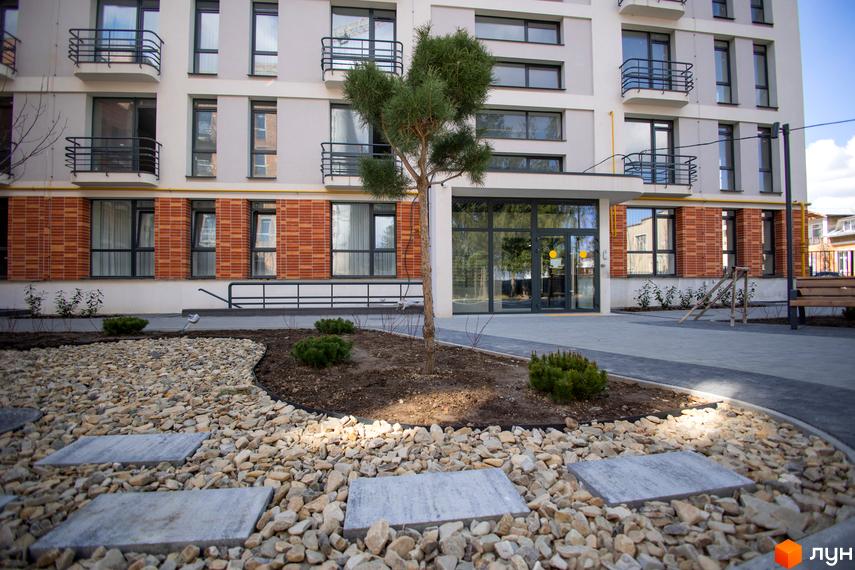 Зовнішній вигляд житлового комплексу РЕМ з цегляним фасадом та балконами. Прибудинкова територія з доріжками та зеленими насадженнями.