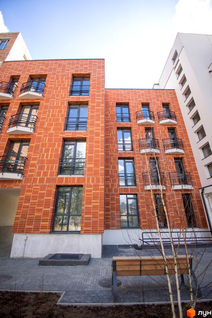 Житловий комплекс РЕМ з фасадом з помаранчевої цегли та балконами. На прибудинковій території бруківка, лавка та молоді дерева.