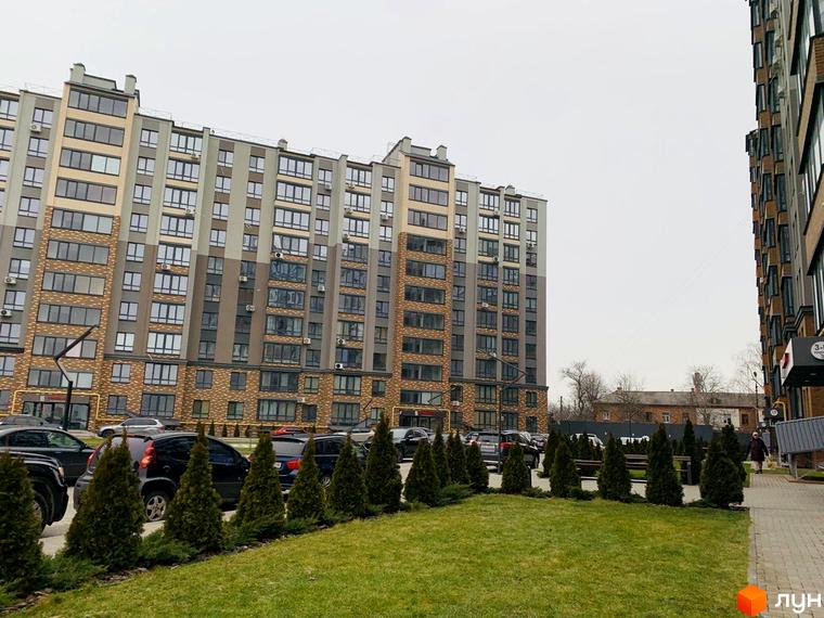 Багатоповерховий будинок ЖК Grand City 2 з великою кількістю вікон. Перед будинком зелена зона з туями та місцями для паркування.