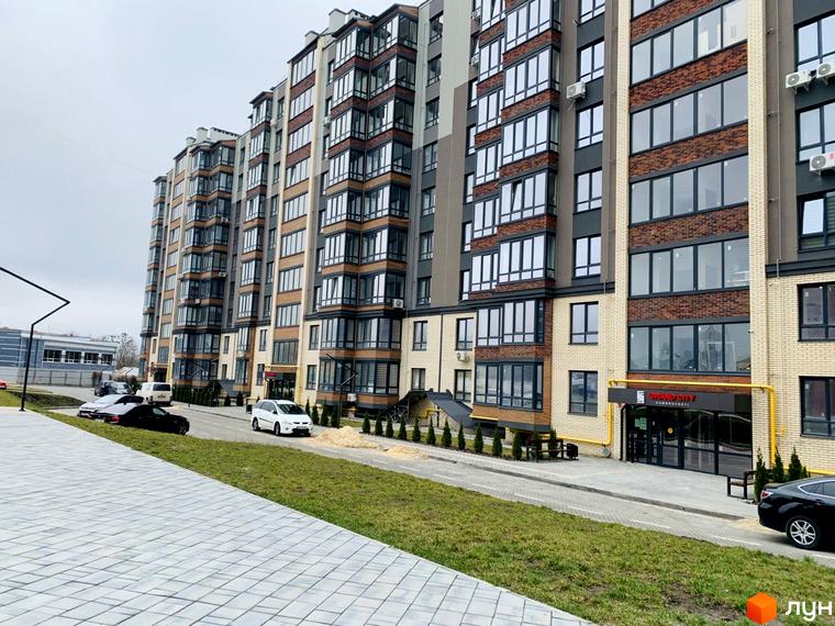 Житловий комплекс Grand City 2 з багатоповерховим будинком, що має цегляні та світлі фасади, великі вікна. Прибудинкова територія включає доріжки, зелені насадження та дорогу з автомобілями.