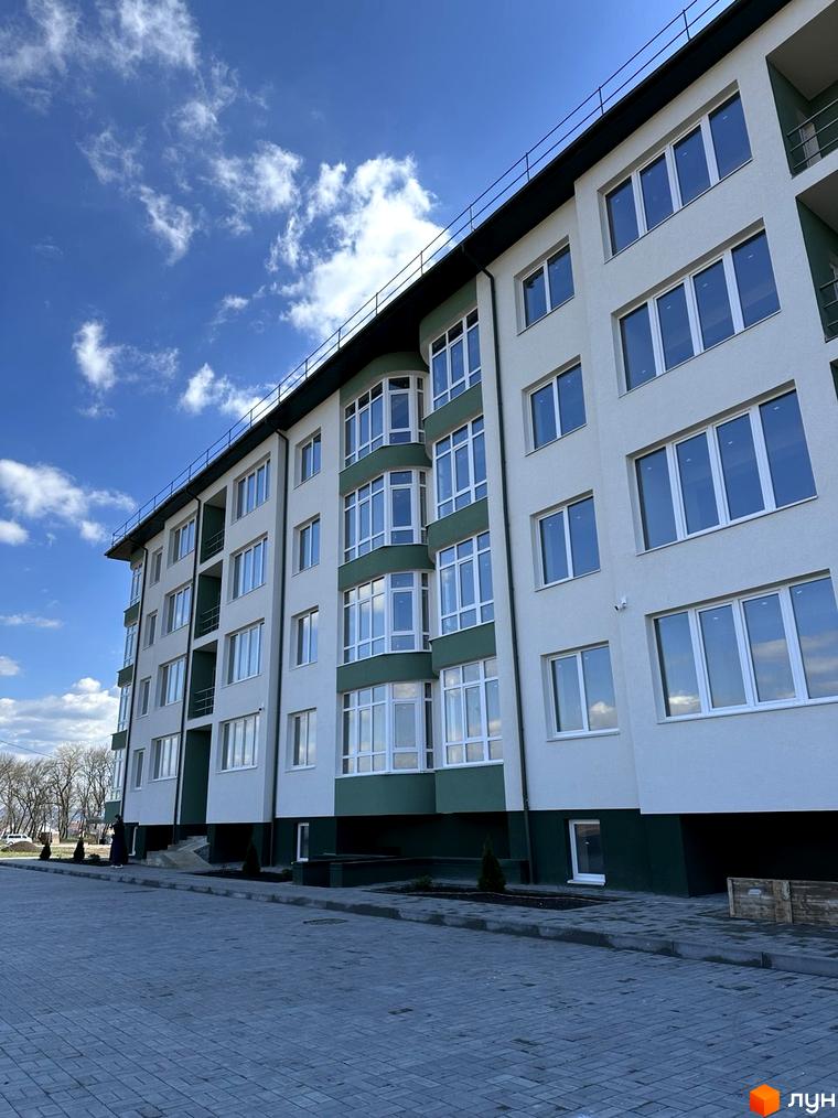 Багатоповерховий будинок з великими вікнами та зеленими вставками на фасаді. Перед будинком викладена бруківка, ростуть невеликі дерева.