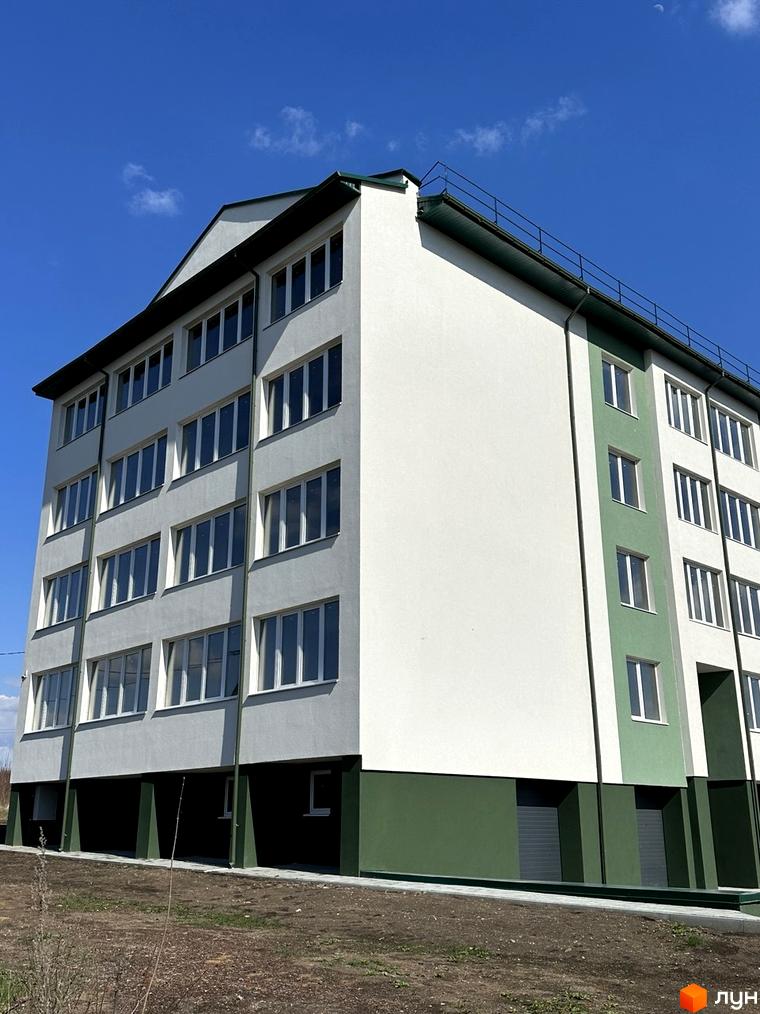 Фасад новобудови білого та зеленого кольорів, з великою кількістю вікон. Навколо будинку видно ґрунт та чисте небо.
