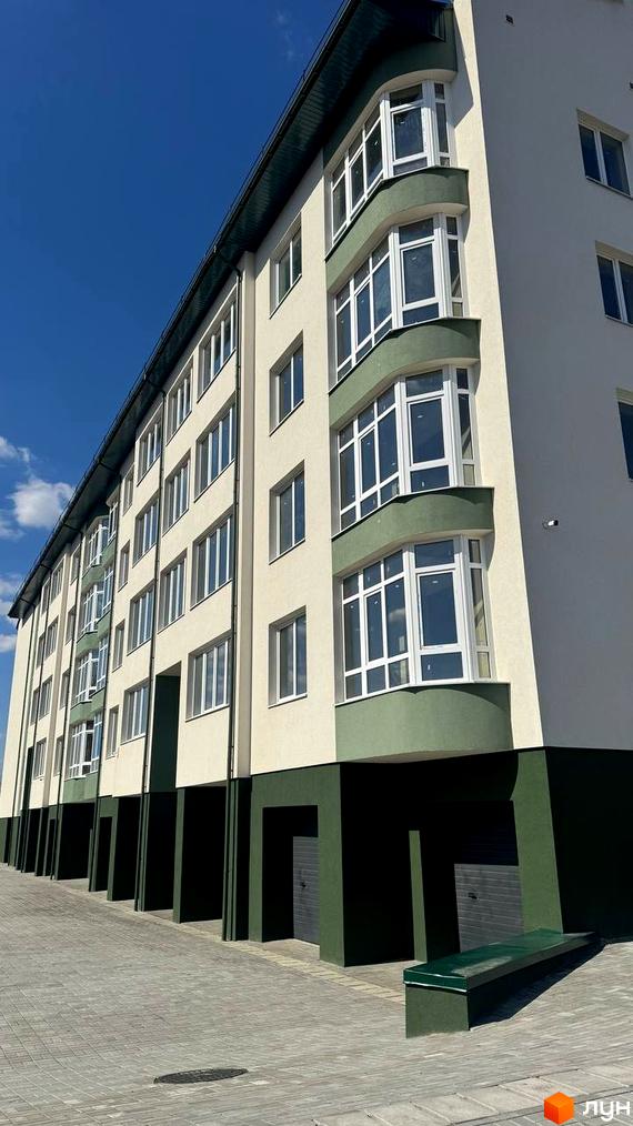 Багатоквартирний будинок світлого кольору з темно-зеленими елементами та еркерами на вул. Спортивна. Перед ним вимощена територія з гаражними воротами.