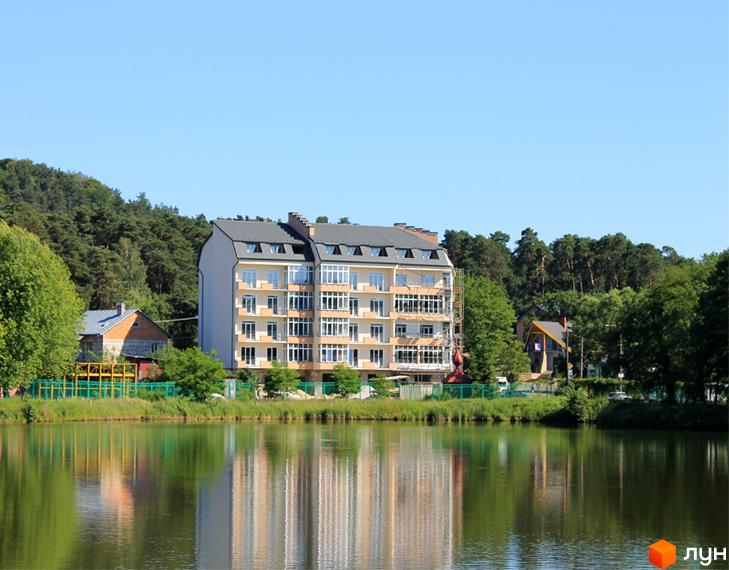 Житловий комплекс Хвиля біля озера. Багатоповерховий будинок світло-бежевого кольору з сірим дахом та численними вікнами. Навколо зелені дерева та трава.