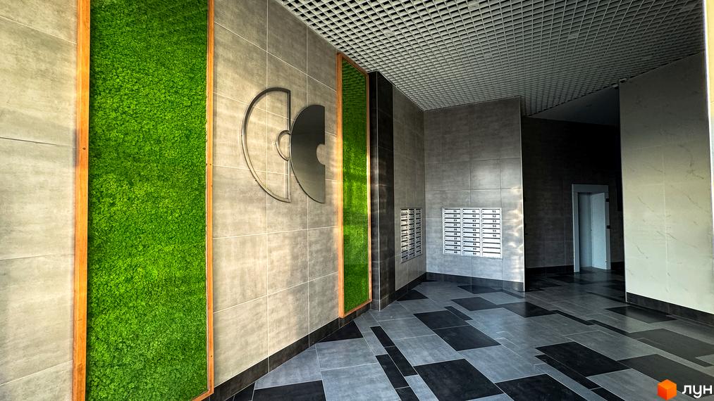 Інтер'єр холу в ЖК Синергія Сіті з декоративною зеленню та логотипом комплексу. Сіра плитка на стінах та підлозі, решітчаста стеля.