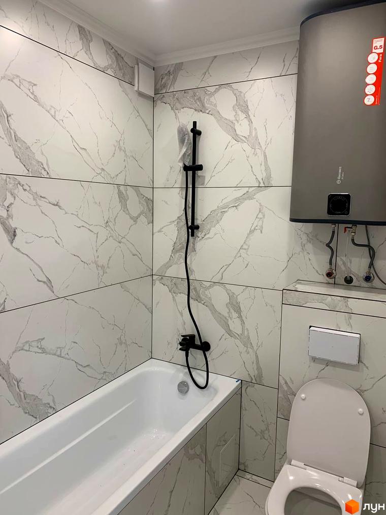 Ванна кімната з білою плиткою під мармур, білою ванною та унітазом. Над унітазом розташований бойлер.