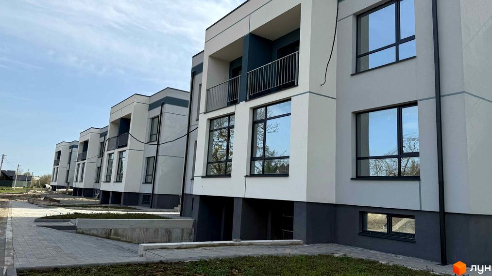 Сучасні таунхауси 4Family House з великими вікнами та балконами на тлі блакитного неба. Перед будинками облаштована прибудинкова територія з бруківкою та зеленими насадженнями.