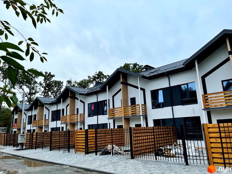 Котеджне містечко Київське: ряд будинків з білими фасадами, чорними дахами та дерев'яними балконами. Вимощена доріжка та дерев'яний паркан.