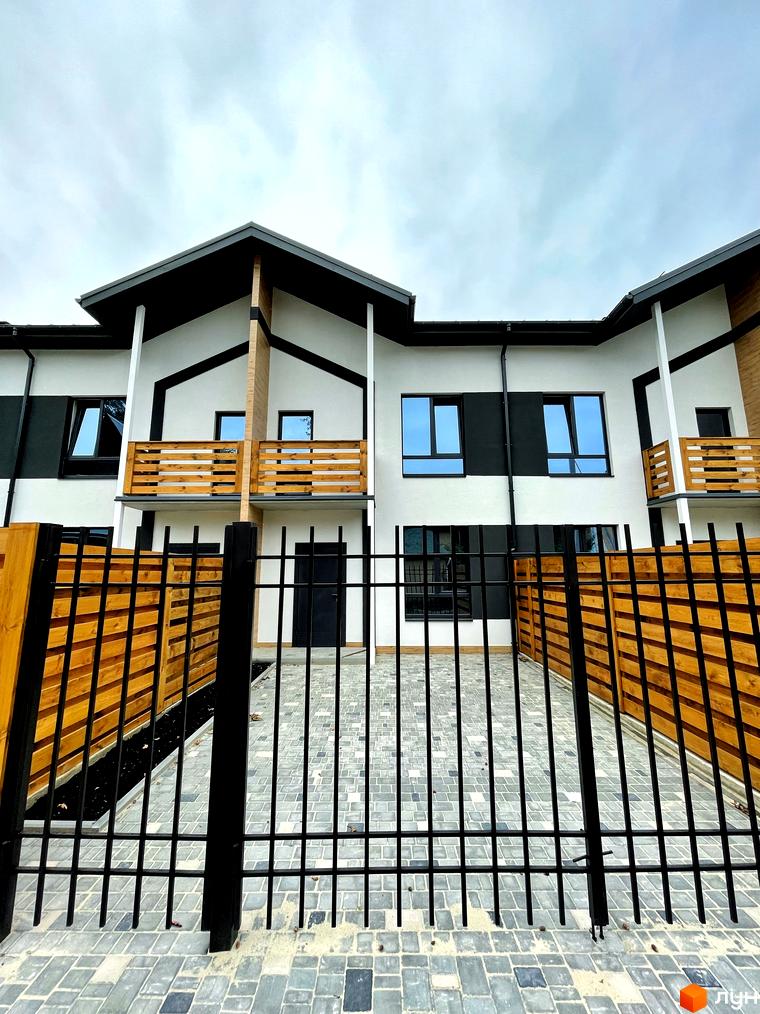 Котеджне містечко Київське: фасади будинків білого кольору з темними дахами та дерев'яними балконами. Прибудинкова територія з бруківкою та металевою огорожею.