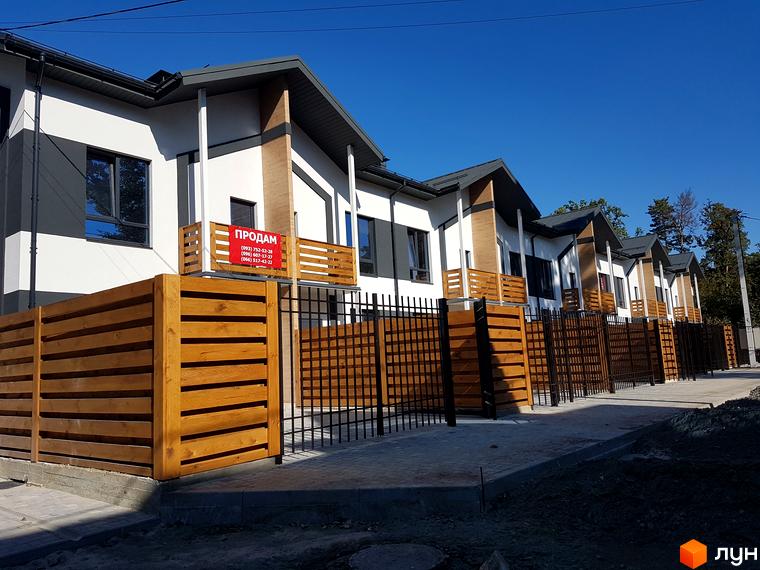 КМ Київське: ряд двоповерхових будинків з білими фасадами, темними дахами та дерев'яними акцентами. Територія огороджена комбінованими парканами.