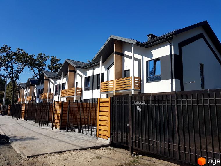 Зовнішній вигляд будинків у Котеджному містечку Київське з білими фасадами, дерев'яними балконами та парканом. Благоустрій території з бруківкою.
