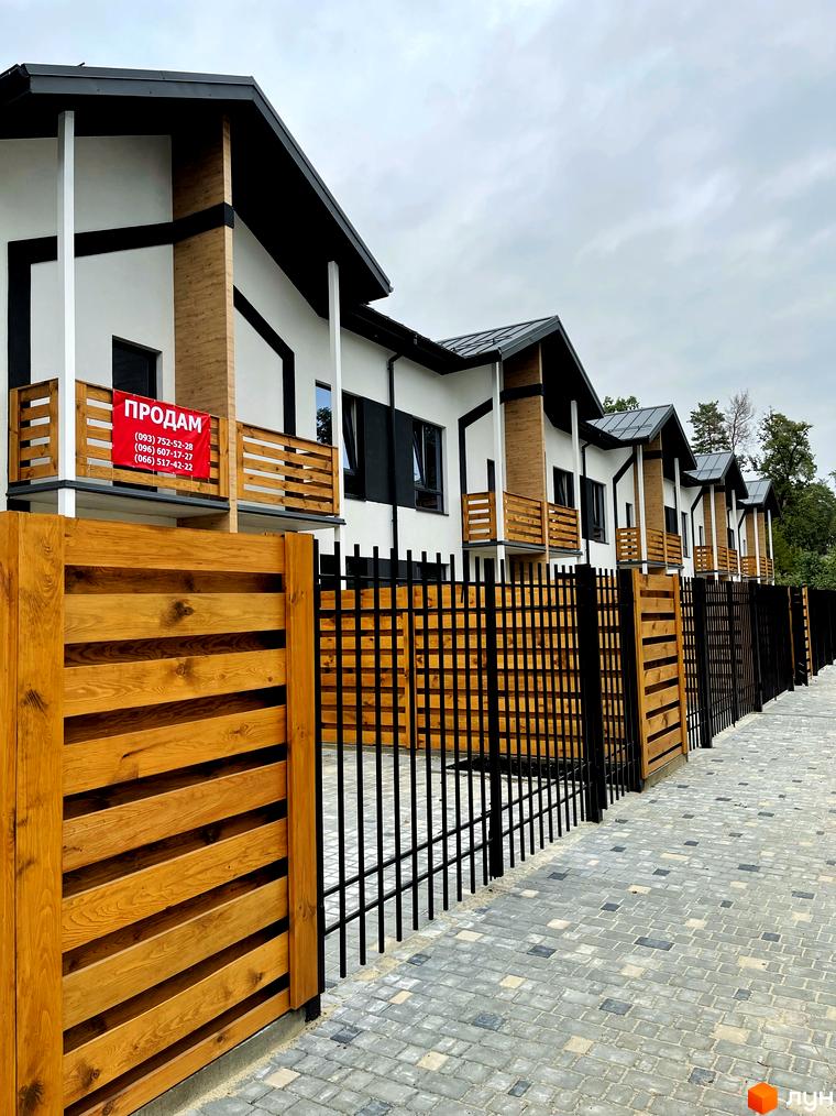 Котеджне містечко Київське: ряди білих будинків з чорними дахами та дерев'яними балконами. Прибудинкова територія з огорожею та бруківкою.