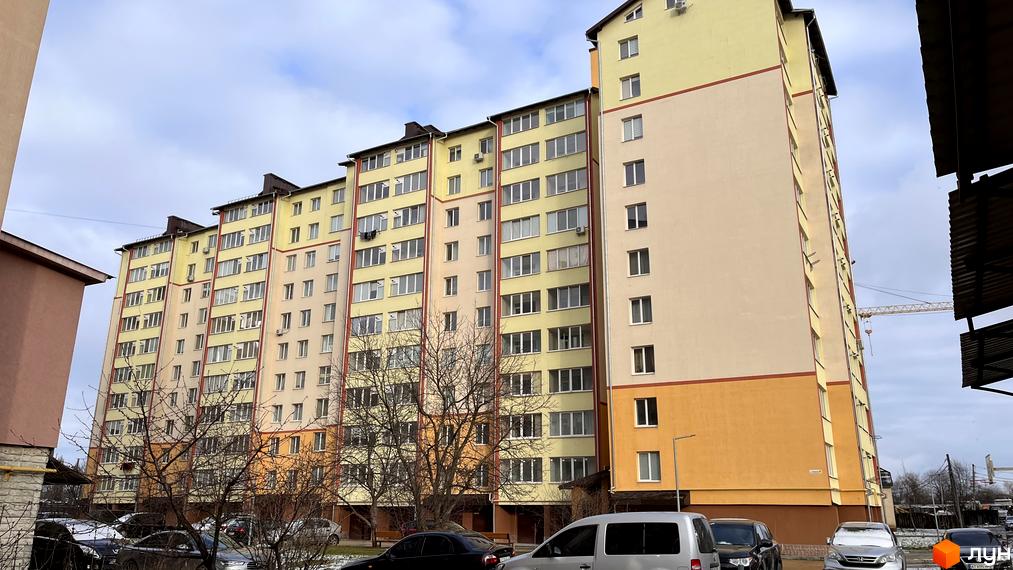 Багатоповерховий будинок з жовтими, бежевими та помаранчевими секціями. На території перед будинком припарковані автомобілі, ростуть дерева.