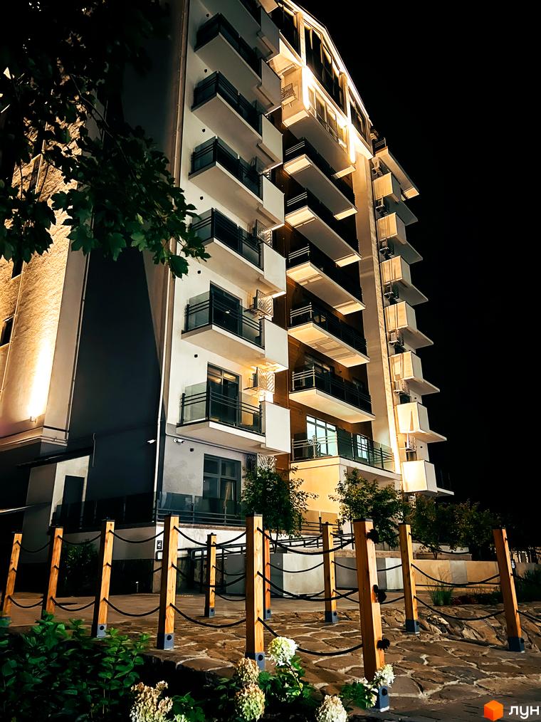 Висотна будівля житлового комплексу з балконами та терасами, освітлена вночі. На передньому плані - огороджена територія з рослинами та декоративними стовпчиками.