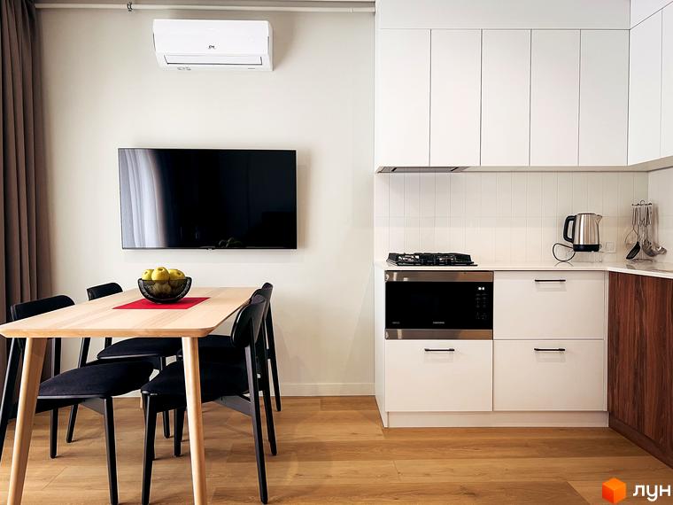 Світла кухня з дерев'яними меблями та телевізором на стіні в апартаментах житлового комплексу Крайобраз. Простора обідня зона з чорними стільцями.
